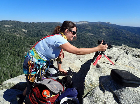 Shira, wearing a climbing harness, with a camera on top of a rock formation.